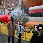 Two students bundled up in winter clothes stand in front of a third holding a camera with balloons from the Macy's Day Parade blown up on the ground next to them