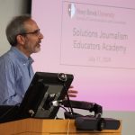 a man delivers a lecture in front of a podium and a powerpoint presentation during the Solutions Journalism Educators Academy