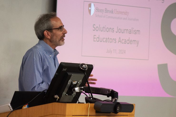a man delivers a lecture in front of a podium and a powerpoint presentation during the Solutions Journalism Educators Academy