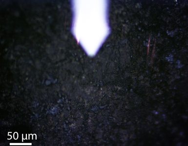 Photo caption: Bennu sample OREX-800066-3 under near-field optical microscope during nanoscale infrared analysis. The metallic tip (top center) of the scanning near-field optical microscope probes the sample's chemical composition at ~20-nanometer resolution, revealing distinct organic-mineral domains within this Bennu fragment returned to Earth by NASA's OSIRIS-REx mission in September 2023. Photo Credit: Stony Brook University and Lawrence Berkeley National Laboratory.