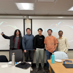 Five students stand in a line in front of projectors, smiling for the camera.