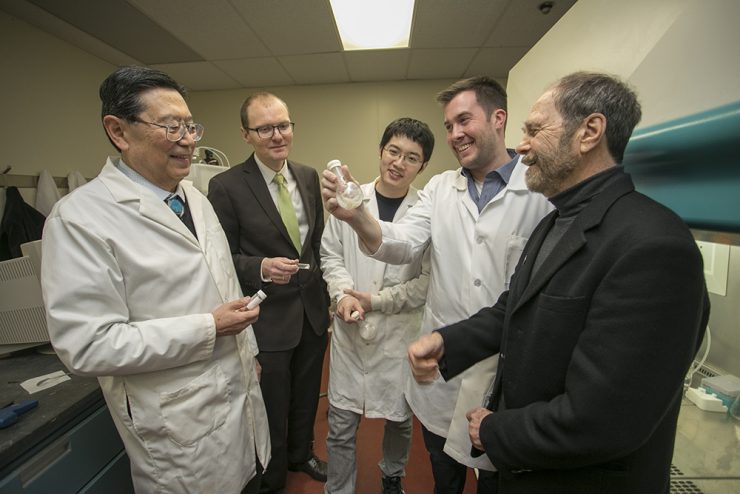 Group of scientists in lab coats gather around as one man holds up a small transparent sample for discussion.