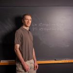 Man in a brown polo stands at a chalkboard with handwritten equations in a classroom setting.